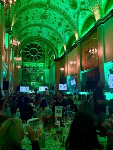 Guests seated at round tables in a high, vaulted room lit in green during the Herd under the Hammer auction for Thames Hospice